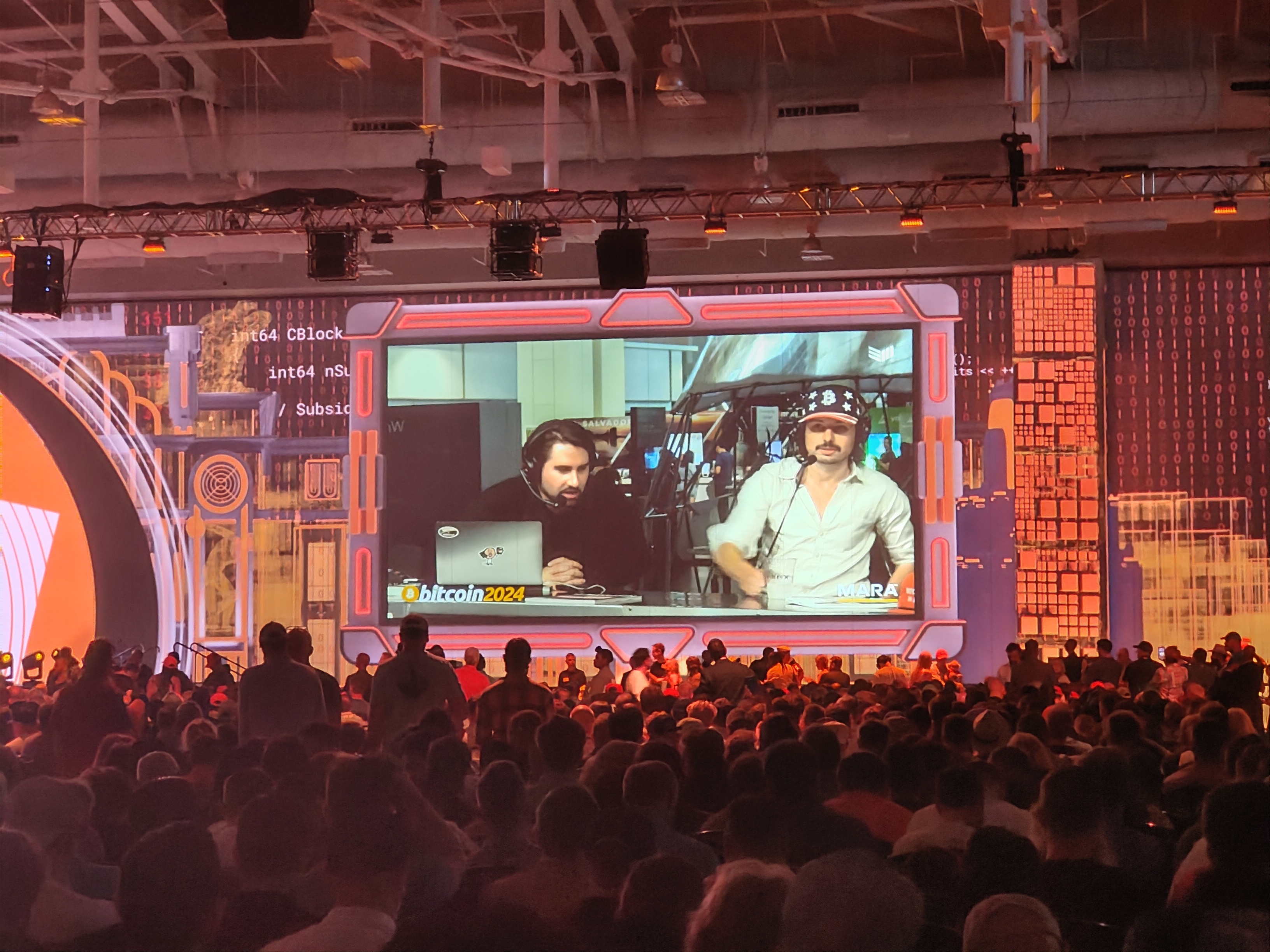 An interview on a big screen. A man is wearing a silly BTC/American flag hat