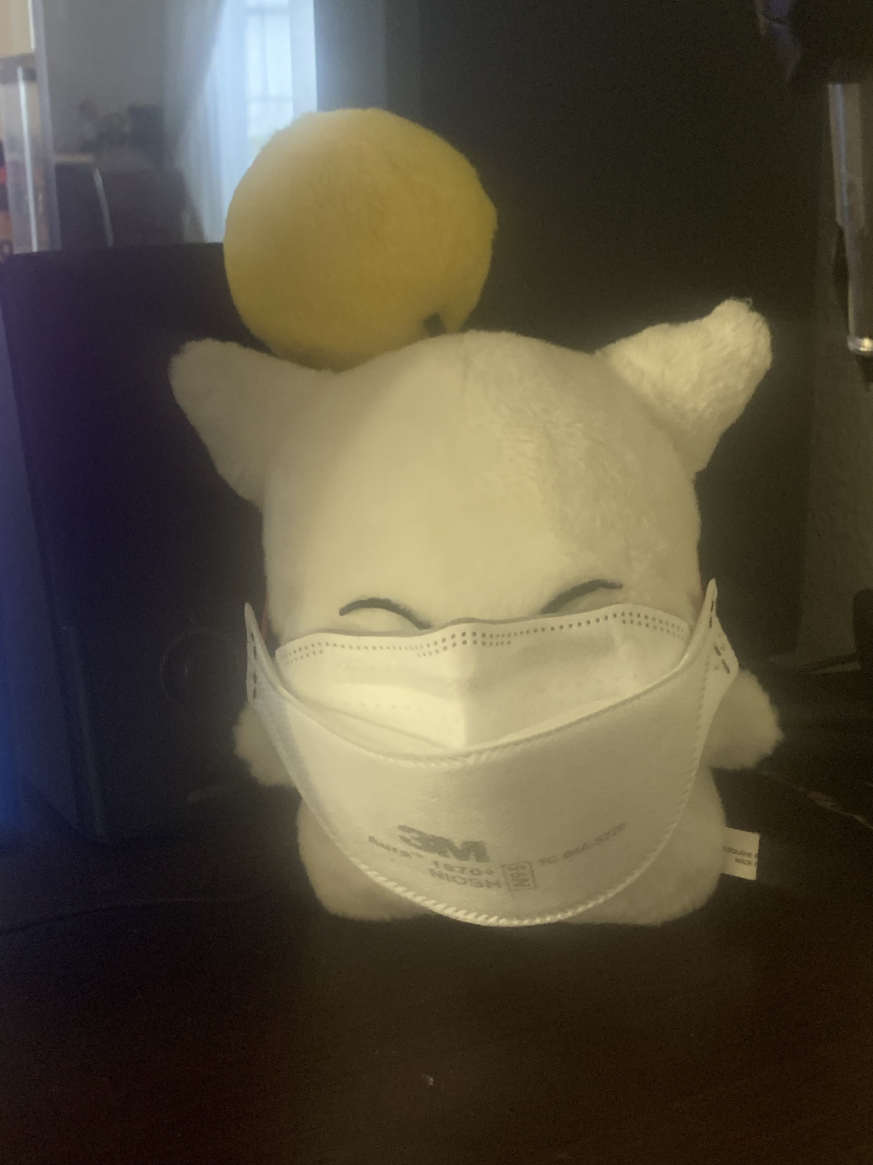 A plush Final Fantasy moogle wearing a human-sized N95 respirator. The mask is too big for it and nearly covers its entire body, with only its small, squinted eyes just visible above the nose ridge.