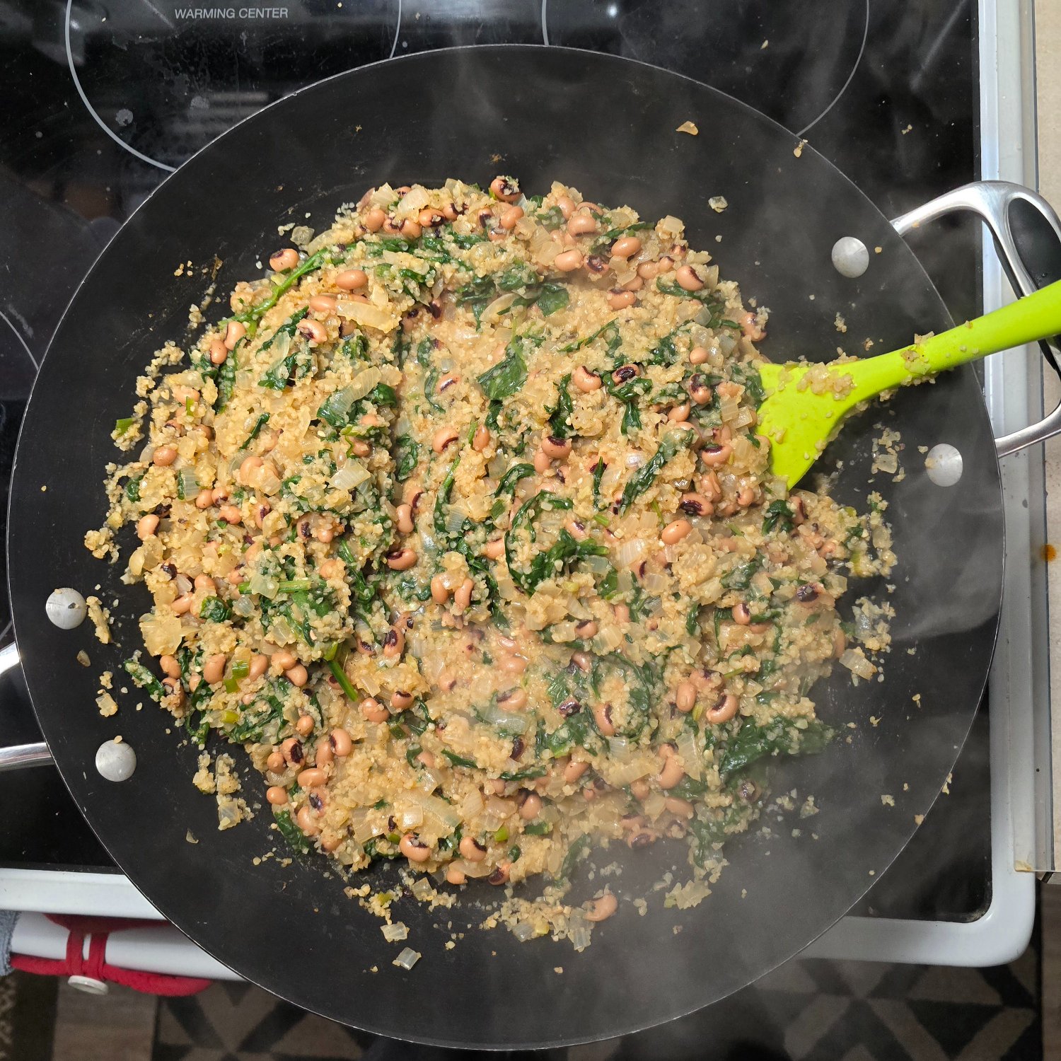 A top down photo of a wok filled with bulgar, onions, garlic, green onions, spinach, black eyed peas, vege broth, baharat spice blend, and lemon juice.  Steam rises into the camera