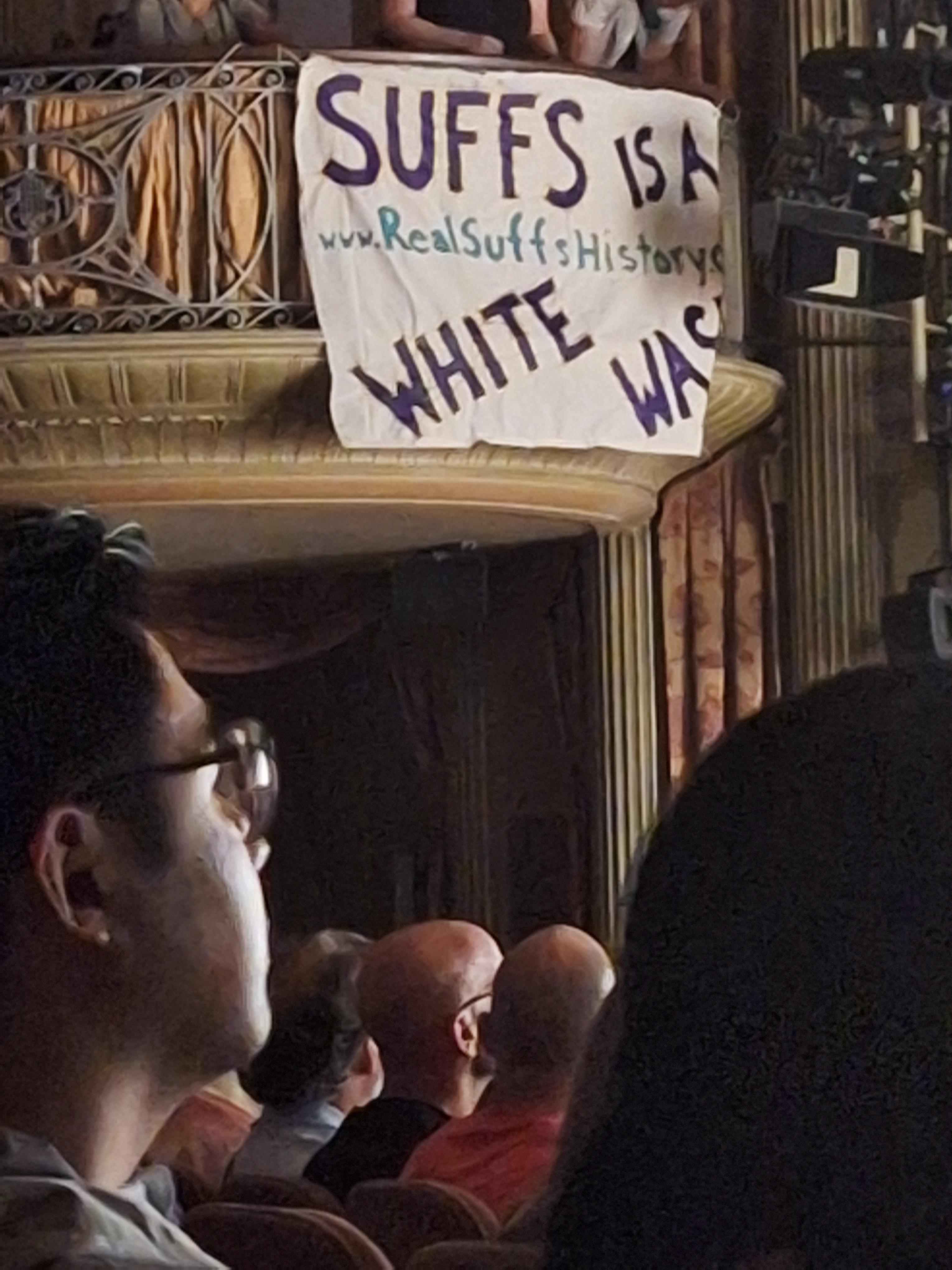 A banner with the message "SUFFS is a white wash" and the website "www.RealSuffsHistories.com" is hung from a balcony in a theater.