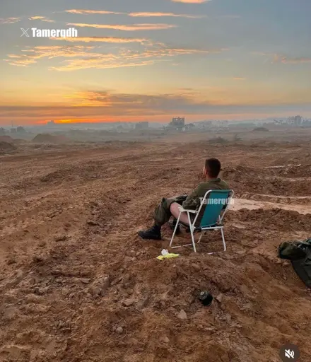 Israeli soldier looking at a destroyed Gaza with their hand inside of their pants, sitting in a folding chair