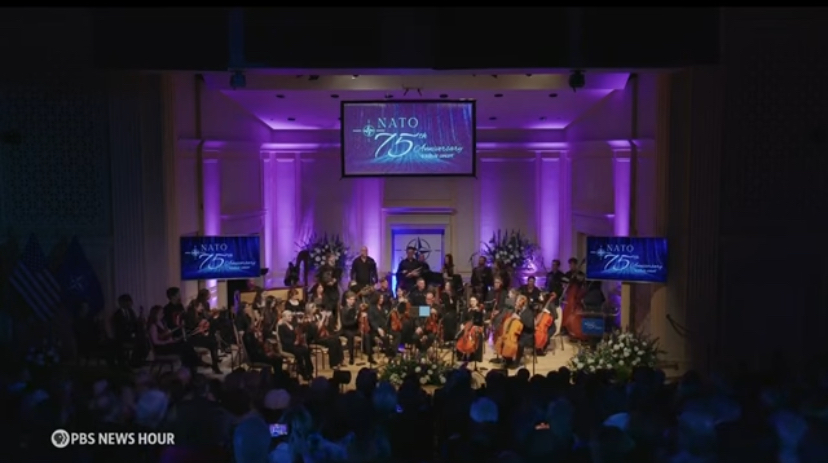 Image showing orchestra playing beneath a large screen reading “NATO 75”