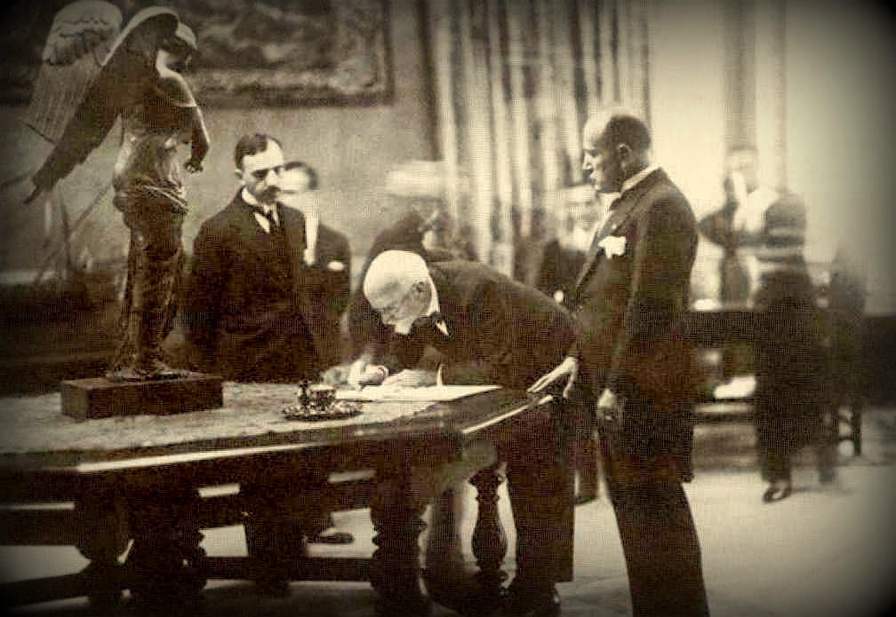 Eleftherios Venizelos hunched over a table to sign this treaty with Fascist Italy. Benito Mussolini stands to his left.