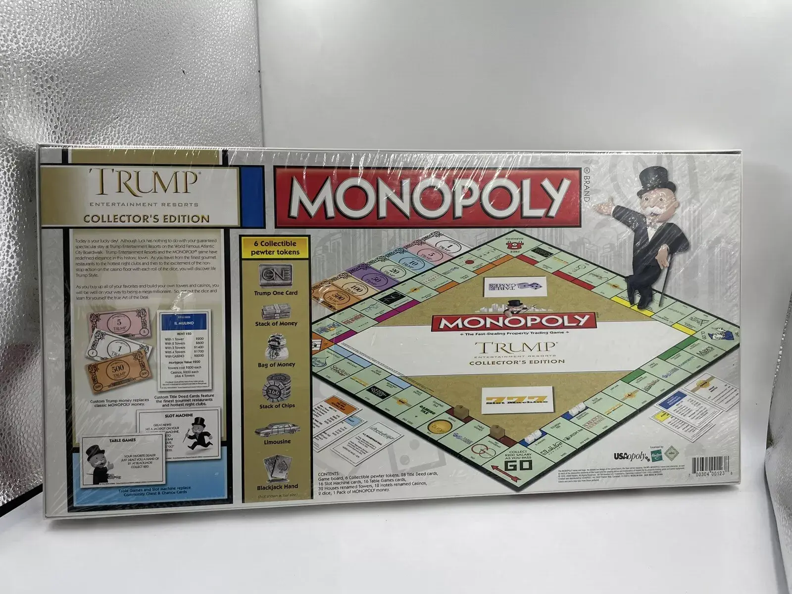 Picture of a real Trump-branded Monopoly board game, back side, highlighting the six unique player pieces. Those pieces include a "Trump ONE credit card", a stack of money, a bag of money, a stack of gambling chips, a limousine, and two cards making a Blackjack (ace of spades showing)